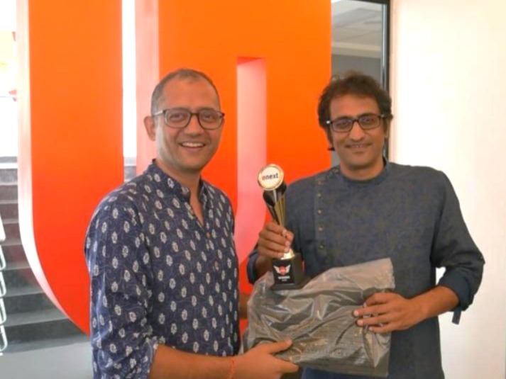 Two smiling men wearing patterned shirts stand indoors. One hands a trophy labeled onest to the other, who also holds a wrapped gift. A large red letter U and CA signage are visible in the background.